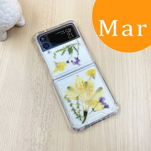 May include: A clear phone case with dried flowers pressed inside. The case is designed for a foldable phone and has a hinge in the middle. The flowers are yellow and purple.