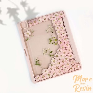 May include: A pink iPad case with a clear back panel featuring pressed pink flowers and greenery. The case has a built-in pencil holder. The text "More Than Resin" is visible below the case.
