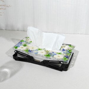 May include: A rectangular tissue box with a floral design. The clear lid is decorated with white, blue, and green flowers. The black base has an adjustable, folding design. White tissues are visible.