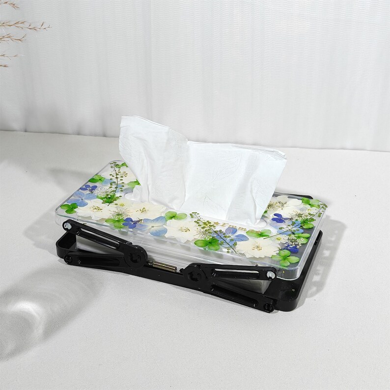 May include: A rectangular tissue box with a floral design. The clear lid is decorated with white, blue, and green flowers. The black base has an adjustable, folding design. White tissues are visible.