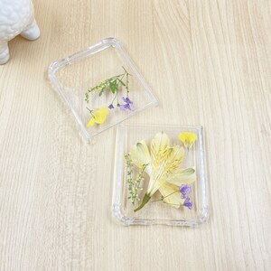 May include: Two clear plastic phone cases with pressed flowers inside. One case has a single yellow flower with purple accents, the other has a larger yellow flower with purple accents.