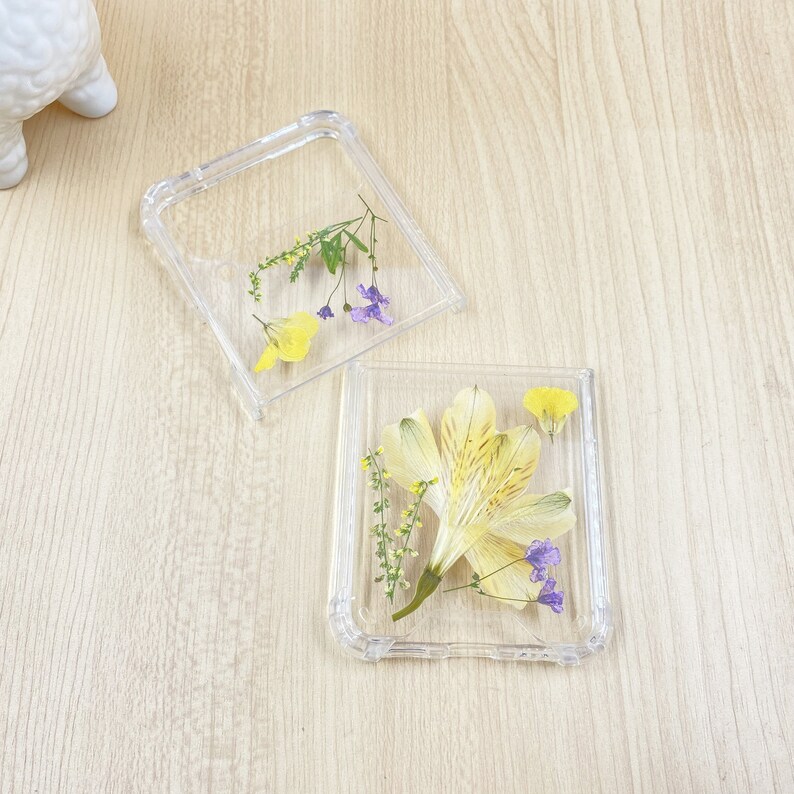May include: Two clear plastic phone cases with pressed flowers inside. One case has a single yellow flower with purple accents, the other has a larger yellow flower with purple accents.