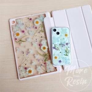 Può includere: Due custodie per telefono con fiori pressati. La custodia a sinistra è una custodia trasparente con fiori bianchi e rosa. La custodia a destra è una custodia trasparente con fiori blu e bianchi. Entrambe le custodie sono su una superficie di legno chiaro.