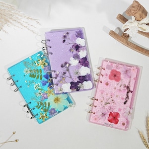May include: Three small, handmade notebooks with floral designs. One is turquoise with yellow and purple flowers, another is lavender with white and purple flowers, and the third is pink with red flowers. Each has a silver spiral binding.