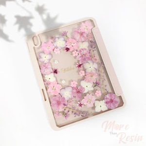 Könnte beinhalten: Eine rosa iPad-Hülle mit Blumenmuster. Die Hülle hat eine transparente Rückseite und einen rosa Rand. Die Blumen sind gepresst und in Harz versiegelt. Der Text "Fraiche" ist in goldenen Buchstaben auf der Hülle geschrieben.