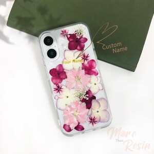 Puede incluir: Una funda transparente para teléfono con un diseño floral. Las flores son rosas, blancas y moradas. La funda tiene una inscripción dorada que dice "Your Name" y "Custom Name".