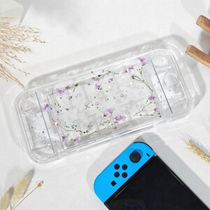 May include: Transparent Switch case decorated with a floral design. The case has a wreath of purple flowers and the name "K a i a" in white. A blue and black Switch controller is visible at the bottom.