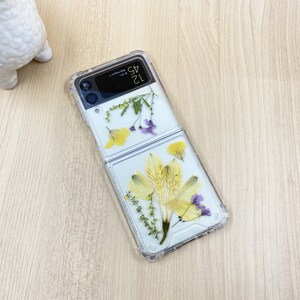 May include: A clear phone case with dried flowers pressed inside. The case is designed for a foldable phone and has a clear back and front. The flowers are yellow and purple.