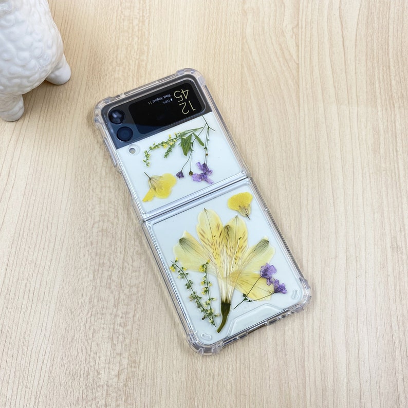 May include: A clear phone case with dried flowers pressed inside. The case is designed for a foldable phone and has a clear back and front. The flowers are yellow and purple.