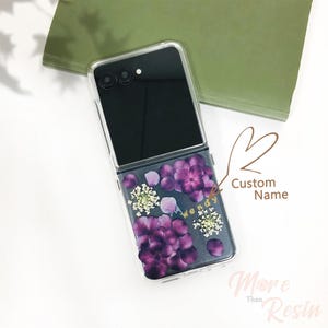 May include: A clear phone case with a floral design. The case has a black background with purple and white flowers pressed into the resin. The case is designed for a foldable phone and has a hinge in the middle. The text "Wendy" is written in gold lettering on the case.