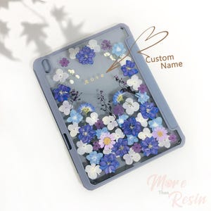 May include: A light blue iPad case with a clear back panel featuring pressed flowers in shades of blue, purple, and white. The text "Rose" is written in gold lettering on the case. The case is designed for a tablet with a screen size of 10.2 inches.