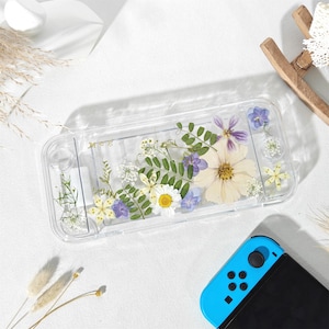 Könnte beinhalten: Transparente Nintendo Switch-Hülle mit getrockneten Blumen in Weiß, Gelb und Lila. Die Hülle hat eine rechteckige Form mit abgerundeten Ecken und dem Wort "Mee". Ein blauer Controller ist sichtbar. Ca. 17 cm lang.