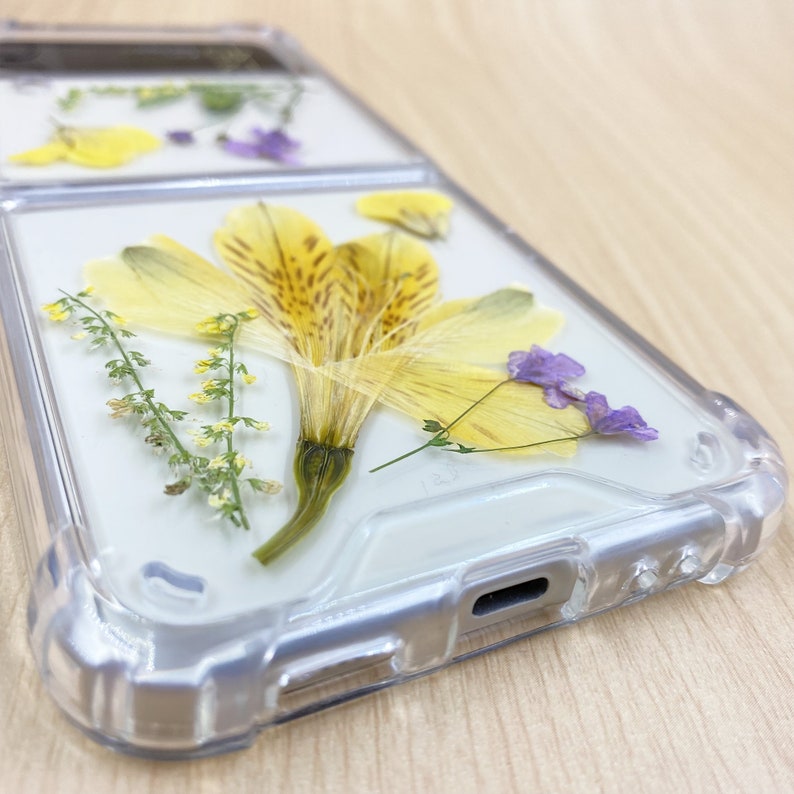 May include: A clear phone case with pressed yellow and purple flowers. The case has a raised edge around the camera lens and a cutout for the charging port.