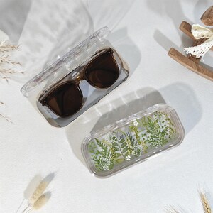 Peut inclure: Lunettes de soleil marron dans un &eacute;tui transparent avec un chiffon de nettoyage gris. Un deuxi&egrave;me &eacute;tui transparent est d&eacute;cor&eacute; de fleurs et de verdure press&eacute;es. Les &eacute;tuis sont rectangulaires et ont un couvercle transparent.