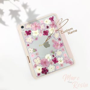 Könnte beinhalten: Eine rosa iPad-Hülle mit einer durchsichtigen Rückseite, die mit gepressten Blumen in Rosa-, Weiß- und Lilatönen verziert ist. Die Hülle hat Platz für einen individuellen Namen, der in brauner Schrift steht. Der Text "More Than Resin" ist in weißer Schrift auf rosa Hintergrund geschrieben.