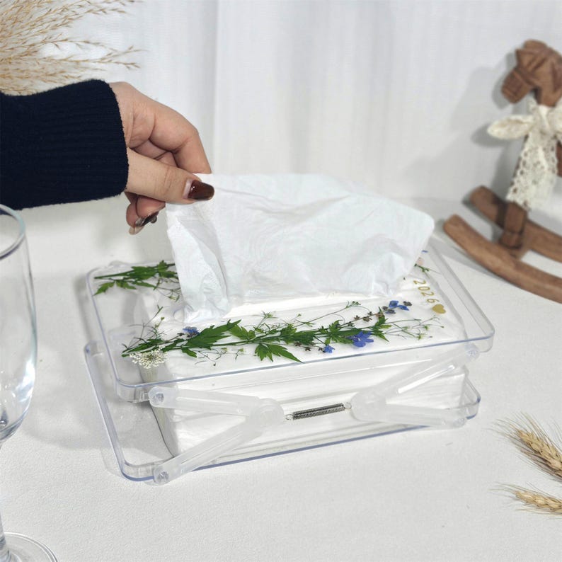 Może przedstawiać: Przezroczyste, prostokątne pudełko na chusteczki z klapką. Pudełko jest ozdobione prasowanymi kwiatami i zielenią. Białe chusteczki są wyciąganie z g&oacute;ry. W tle dekoracyjny drewniany konik na biegunach.