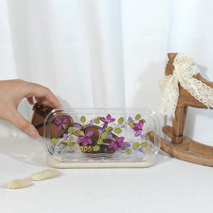 Peut inclure: Un r&eacute;cipient rectangulaire transparent avec un motif floral, contenant des chocolats en forme de c&oelig;ur. Le r&eacute;cipient a une base beige et un couvercle transparent d&eacute;cor&eacute; de fleurs violettes et de feuilles vertes. Le texte "Good Luck 005" est visible.