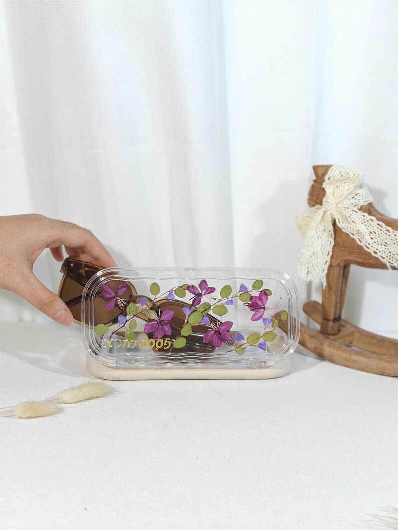 Peut inclure: Un r&eacute;cipient rectangulaire transparent avec un motif floral, contenant des chocolats en forme de c&oelig;ur. Le r&eacute;cipient a une base beige et un couvercle transparent d&eacute;cor&eacute; de fleurs violettes et de feuilles vertes. Le texte "Good Luck 005" est visible.
