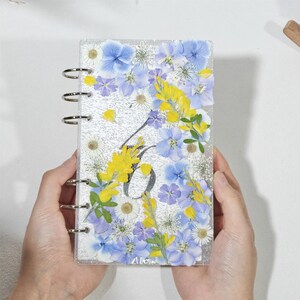 Op de afbeelding: Een helder, rechthoekig notitieboek met zilveren metalen ringen, gevuld met geperste blauwe, gele en witte bloemen. Het notitieboek heeft een glitterachtergrond en de letter "A" is zichtbaar. Het notitieboek wordt door twee handen vastgehouden.