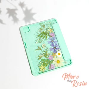 May include: A mint green tablet case featuring a clear back panel embedded with an arrangement of pressed dried flowers, including purple, white, and yellow blossoms with green foliage. The text More Than Resin is printed in the bottom right corner.
