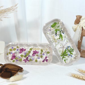 Peut inclure: Deux bo&icirc;tes rectangulaires transparentes avec des fleurs press&eacute;es et l'inscription "GOOD LUCK" en lettres dor&eacute;es. L'une des bo&icirc;tes est droite, l'autre est couch&eacute;e. Les fleurs sont violettes, blanches et vertes.