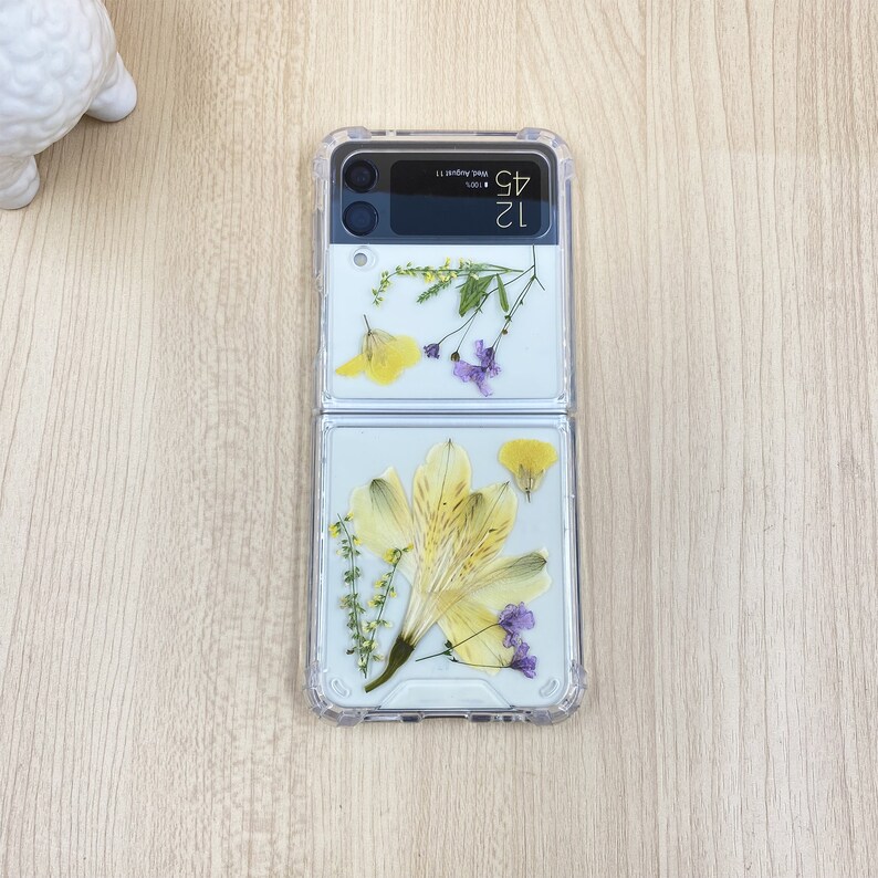 May include: A clear phone case with a floral design. The case is made of two pieces that snap together. The top piece has a yellow flower and purple flowers. The bottom piece has a larger yellow flower and a few purple flowers. The phone case has a digital clock that reads "12:43" and the text "Wed, August 17".