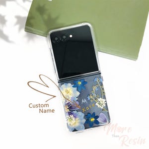 May include: A clear phone case with a floral design. The case is for a foldable phone and has a black back. The flowers are blue, white, and purple. The case has a gold inscription that says "Kate".