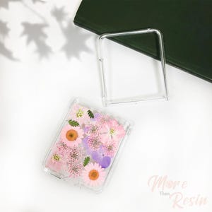May include: A clear phone case with a floral design. The case is filled with pressed pink, purple, and white flowers.
