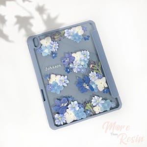 May include: A light blue iPad case with a floral design featuring pressed blue and white flowers. The case has a clear back and a solid blue border. The name "Johnson" is printed on the case.