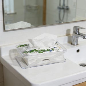 Może przedstawiać: Przezroczysty plastikowy pojemnik na chusteczki z kwiatowym wzorem na pokrywie, zawierający białe chusteczki. Pudełko stoi na białym blacie w pobliżu umywalki i lustra.