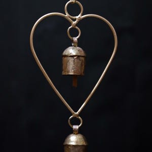 May include: A heart-shaped wind chime with two small bells. The metal frame is a light gold color, and the bells have a rustic, aged appearance. The background is a solid black.
