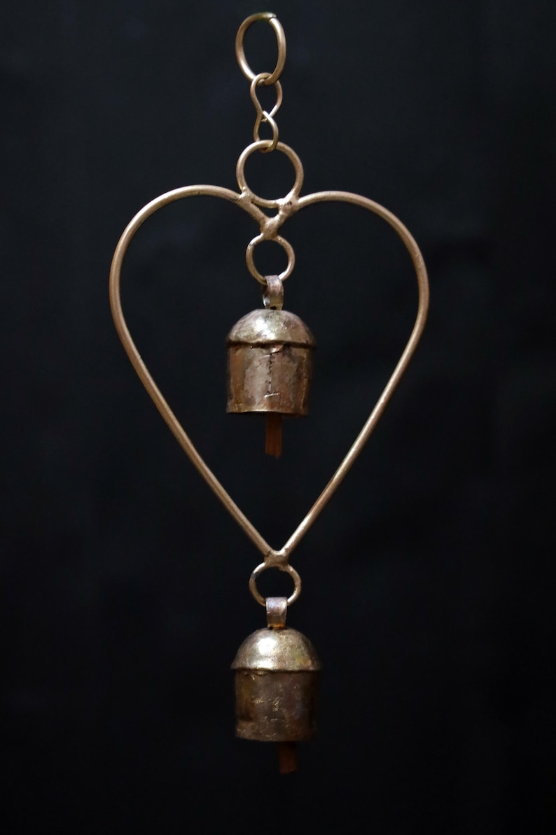 May include: A heart-shaped wind chime with two small bells. The metal frame is a light gold color, and the bells have a rustic, aged appearance. The background is a solid black.
