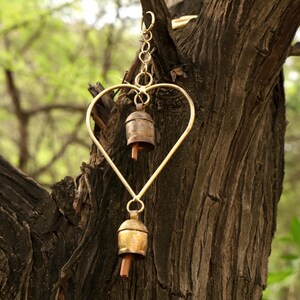 May include: A heart-shaped wind chime with two small bells hanging from a gold-colored metal frame. The bells have a rustic, aged appearance. The chime is suspended from a tree branch against a blurred green background.