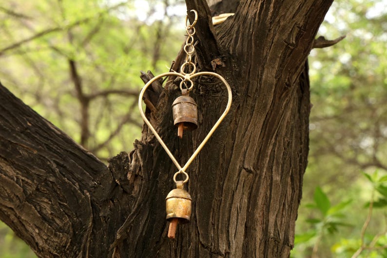 May include: A heart-shaped wind chime with two small bells hanging from a gold-colored metal frame. The bells have a rustic, aged appearance. The chime is suspended from a tree branch against a blurred green background.