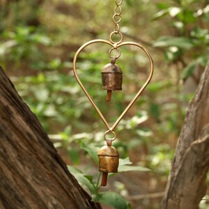 May include: A heart-shaped wind chime with two small bells. The heart is made of gold-colored metal, and the bells are bronze. The background is a blurred view of green foliage and tree trunks.