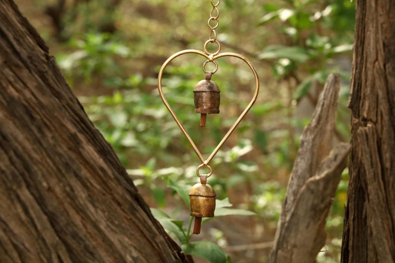 May include: A heart-shaped wind chime with two small bells. The heart is made of gold-colored metal, and the bells are bronze. The background is a blurred view of green foliage and tree trunks.