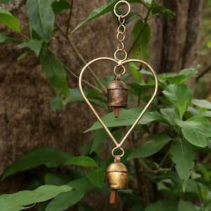 May include: A heart-shaped wind chime with two bells. The chime is made of gold-colored metal and hangs from a chain. The bells are a bronze color. The background is a mix of green leaves and a brown rock.
