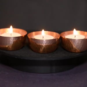 May include: Three copper-colored candle holders with lit white candles. The holders have a hammered texture and sit on a black, round tray. The flames are bright against a dark background.