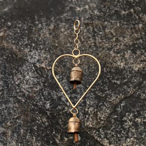 May include: A heart-shaped wind chime with two small bells. The chime is made of gold-colored metal, with a heart-shaped frame and a chain for hanging. The bells have a rustic, aged appearance.