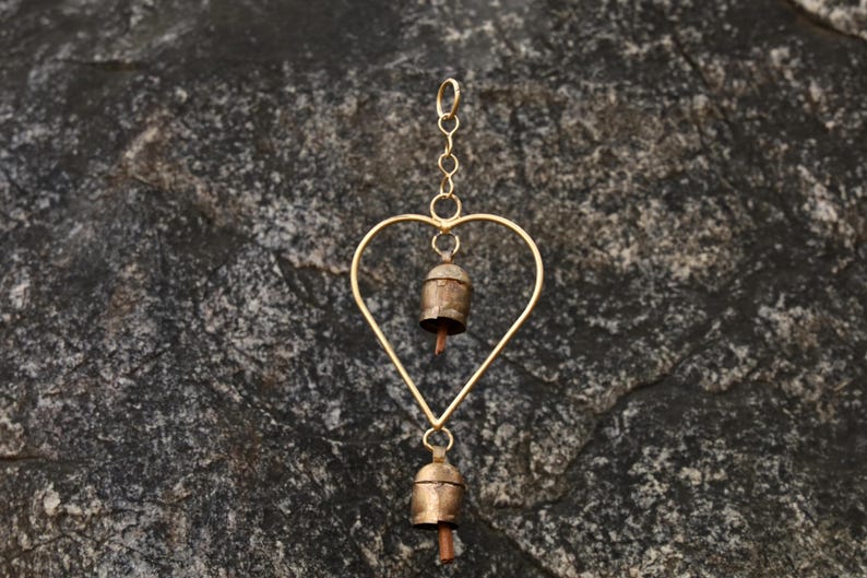 May include: A heart-shaped wind chime with two small bells. The chime is made of gold-colored metal, with a heart-shaped frame and a chain for hanging. The bells have a rustic, aged appearance.