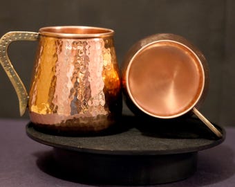 May include: Two copper mugs with hammered textures and brass handles. One mug is upright, the other is on its side, revealing the interior. The mugs sit on a black surface, with a dark background.