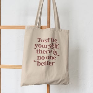 May include: A beige canvas tote bag with brown straps. The bag features a red graphic that reads "Just be yourself, there is no one better."