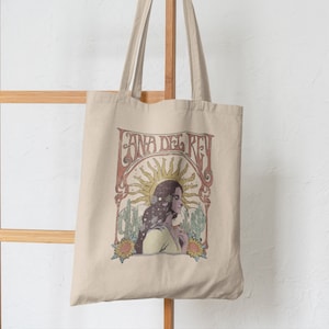 May include: A beige tote bag with a vintage-style graphic of Lana Del Rey. The graphic features a woman with long dark hair, a sun, and cacti. The text "Lana Del Rey" is in a retro font.