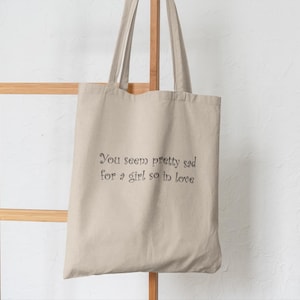 May include: A beige tote bag with long handles. The bag displays the text "You seem pretty sad for a girl so in love" in a dark, elegant font. The bag is made of a light-coloured fabric and hangs against a light-coloured background.