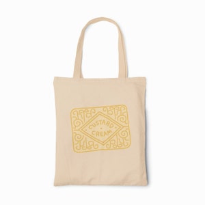 May include: A beige tote bag featuring a yellow outline of a custard cream biscuit. The biscuit has the words "Custard Cream" printed on it.