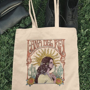 May include: A beige tote bag with a vintage-style graphic of Lana Del Rey. The graphic features a woman with long hair, a sun, and cacti. The text "Lana Del Rey" is written in a stylized font.