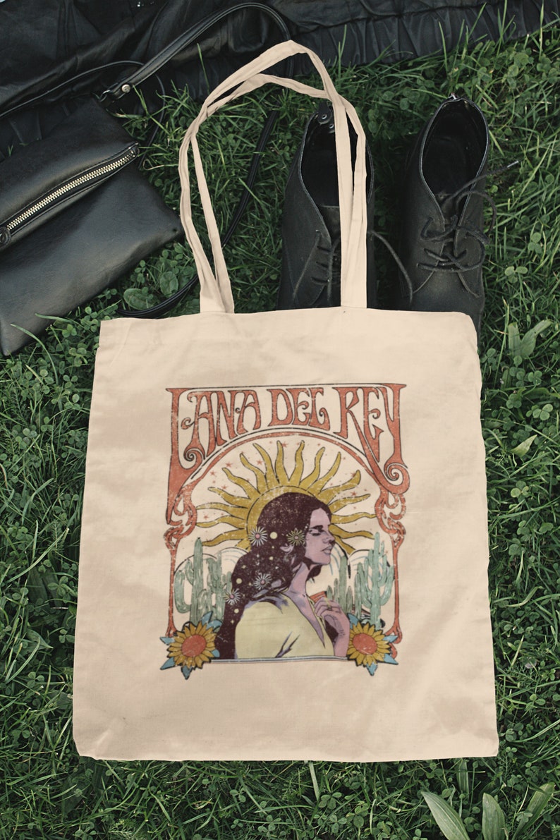 May include: A beige tote bag with a vintage-style graphic of Lana Del Rey. The graphic features a woman with long hair, a sun, and cacti. The text "Lana Del Rey" is written in a stylized font.