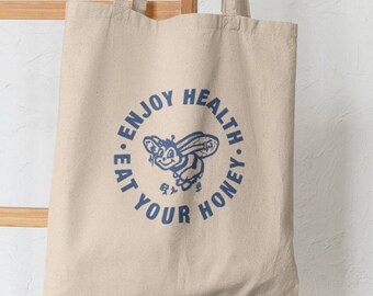 May include: Beige canvas tote bag with a blue circular graphic. The graphic features a cartoon bee and the text "EAT YOUR HONEY. ENJOY HEALTH." The bag has two handles and is made of a sturdy fabric.