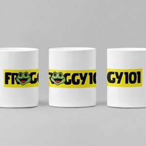 May include: White ceramic mug with a wraparound design featuring the text "FROGGY 101" in black letters on a yellow background. A cartoon frog with a green face and a smiling expression is in the center of the design. The mug has a curved handle.