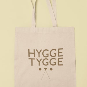 May include: A beige canvas tote bag with brown straps and the text "HYGGE TYGGE" printed on the front. The text is in a bold, sans-serif font.